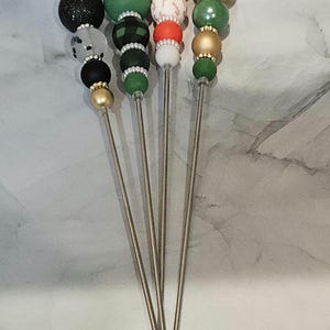 Puede incluir: Juego de cinco brochetas decorativas con adornos de cuentas de colores. Las cuentas son de varios colores, incluyendo negro, verde, rojo y dorado. Cada brocheta tiene un tallo de metal plateado y un diseño de cuentas decorativas.