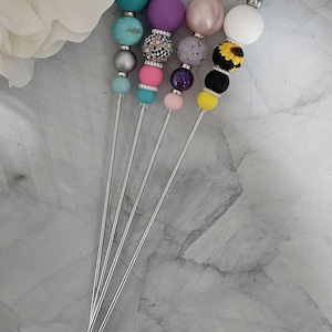 Puede incluir: Un conjunto de brochetas decorativas con coloridas cuentas en la parte superior. Cada brocheta presenta un diseño diferente, incluyendo turquesa, morado, rosa y un patrón de girasol. Las brochetas tienen tallos de metal plateado, ideales para aperitivos o cócteles.