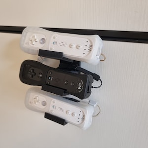 Könnte beinhalten: Drei weiße Wii-Fernbedienungen sind an einer schwarzen Wandhalterung montiert. Die Fernbedienungen sind mit "Wii" beschriftet und haben Tasten und einen Lautsprecher.