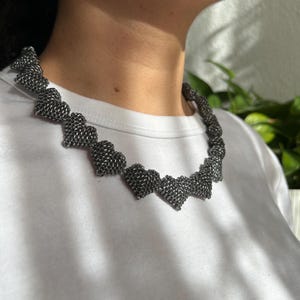 May include: A silver-colored necklace featuring a series of heart-shaped links. The necklace is made of a woven metal material and is worn around the neck. The background includes a white shirt and green foliage.