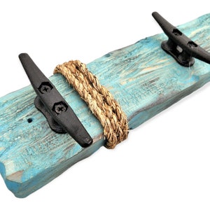 May include: A nautical-themed wall-mounted coat rack. It features a distressed, turquoise-painted wooden plank with three black metal cleats and a natural rope accent. The rustic design evokes a coastal aesthetic.