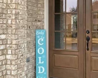 Può includere: Un cartello rettangolare in legno turchese con la scritta "Baby It's Cold Outside" in bianco. Il cartello è decorato con fiocchi di neve ed è posto accanto a un muro di mattoni e a una porta marrone.