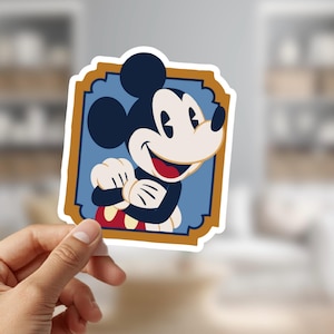 May include: A sticker featuring Mickey Mouse in a classic pose, smiling with arms crossed. The cartoon character is set against a blue background within a decorative gold-coloured frame. The sticker is held by a hand.