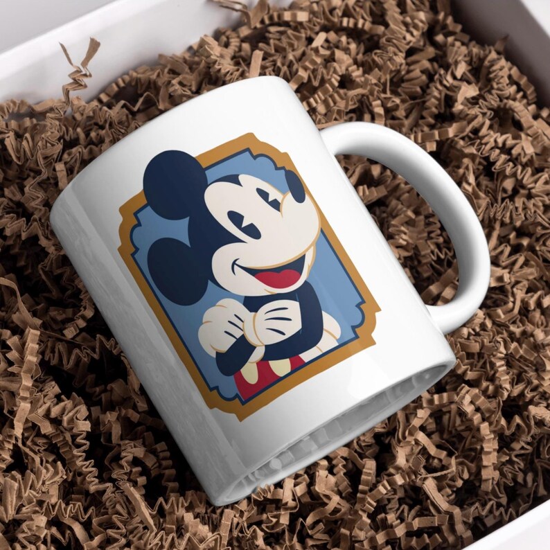 May include: White ceramic mug with a smiling Mickey Mouse graphic. The cartoon character is framed in a blue and gold design. The mug has a white handle and is presented in a white box with brown packing material.