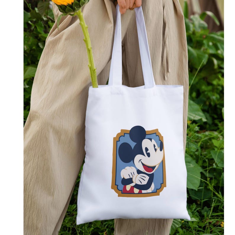 May include: White tote bag with a Mickey Mouse graphic in a blue and gold frame. The bag has white handles. The background shows green foliage and tan trousers.