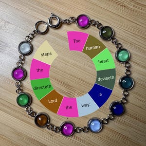May include: A silver bracelet with colorful, round gemstone accents. The bracelet surrounds a circular graphic with text that reads "The human heart deviseth a way:" in a rainbow color scheme. The bracelet is on a wooden surface.