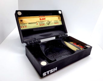 May include: Black rolling tray with a magnetic lid, featuring the text "STSH" and a RAW rolling paper pack inside. The lid has the text "#rawlife RAW NATURAL UNREFINED ROLLING PAPERS" and "The Baller" printed on it.