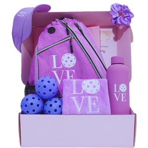 May include: A pink gift box filled with a purple and white "LOVE" themed pickleball set. The set includes a drawstring bag, a towel, four pickleballs, a water bottle, and a purple hair scrunchie.
