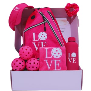 Puede incluir: Una caja de regalo rosa llena de un set de pickleball rosa y blanco con el tema "LOVE". El set incluye una bolsa de pickleball rosa, una toalla rosa, una botella de agua rosa, un gorro rosa, un coletero rosa y cuatro pelotas de pickleball rosas.