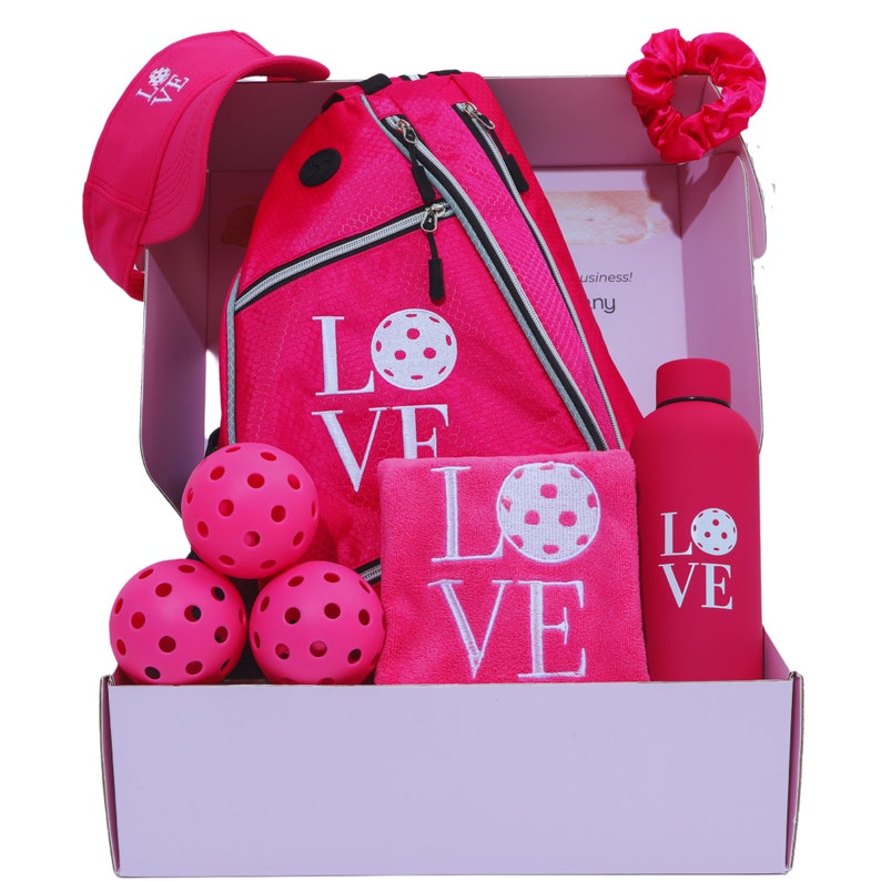 Puede incluir: Una caja de regalo rosa llena de un set de pickleball rosa y blanco con el tema "LOVE". El set incluye una bolsa de pickleball rosa, una toalla rosa, una botella de agua rosa, un gorro rosa, un coletero rosa y cuatro pelotas de pickleball rosas.