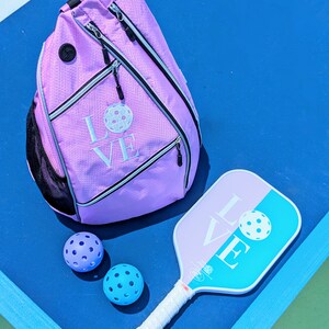 May include: A pink pickleball backpack with the word "LOVE" embroidered on it, along with two blue pickleballs and a blue and pink pickleball paddle.