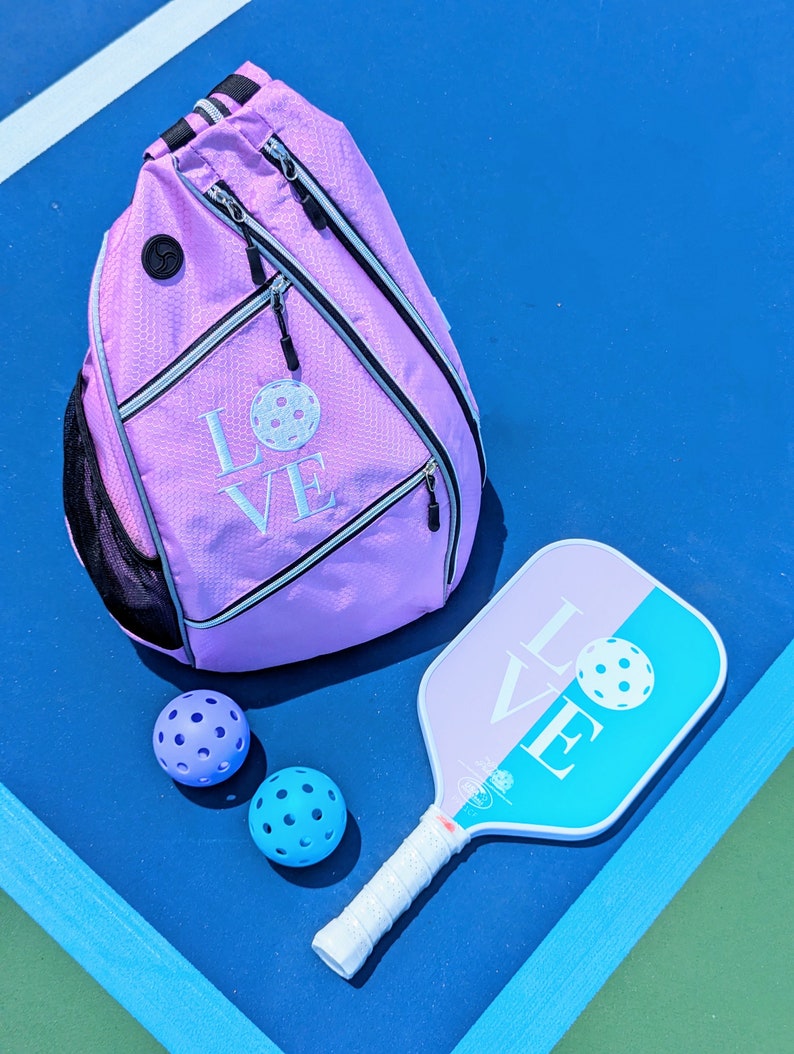 May include: A pink pickleball backpack with the word "LOVE" embroidered on it, along with two blue pickleballs and a blue and pink pickleball paddle.