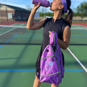 May include: A woman on a pickleball court wearing a black sleeveless shirt, a purple visor, and a purple pickleball bag with the word "LOVE" and a pickleball graphic. She is holding a purple water bottle.