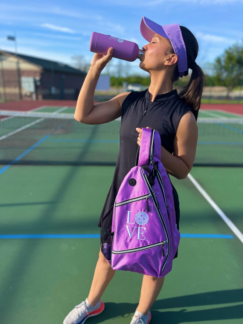 May include: A woman on a pickleball court wearing a black sleeveless shirt, a purple visor, and a purple pickleball bag with the word "LOVE" and a pickleball graphic. She is holding a purple water bottle.