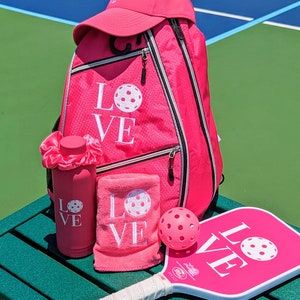 Puede incluir: Conjunto de pickleball rosa con una mochila, una botella de agua, una toalla, una pala y una pelota. La mochila, la toalla, la botella de agua y la pala tienen todas la palabra "LOVE" bordada con un gr&aacute;fico de pickleball.