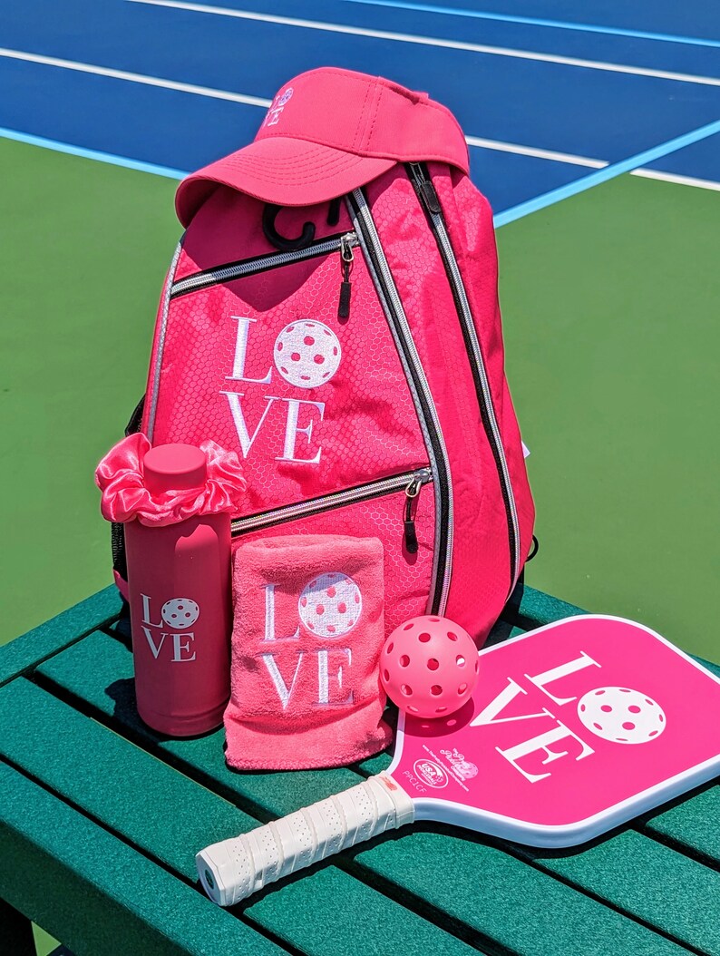 Puede incluir: Conjunto de pickleball rosa con una mochila, una botella de agua, una toalla, una pala y una pelota. La mochila, la toalla, la botella de agua y la pala tienen todas la palabra "LOVE" bordada con un gr&aacute;fico de pickleball.