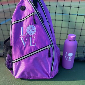 May include: Purple pickleball bag with a white "LOVE" and pickleball graphic embroidered on the front. A matching purple water bottle with the same graphic is next to the bag.