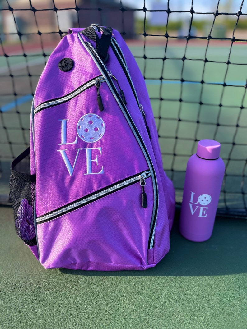 May include: Purple pickleball bag with a white "LOVE" and pickleball graphic embroidered on the front. A matching purple water bottle with the same graphic is next to the bag.