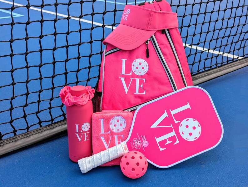 Puede incluir: Raqueta de pickleball rosa, botella de agua rosa, toalla rosa, mochila rosa y visera rosa con la palabra "LOVE" en cada art&iacute;culo. Los art&iacute;culos est&aacute;n en una cancha de pickleball azul.