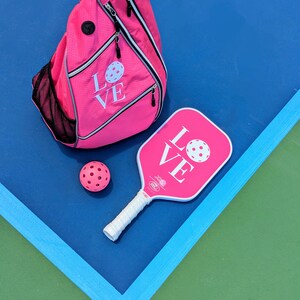Puede incluir: Una pala de pickleball rosa con la palabra "LOVE" impresa en ella, una bolsa de pickleball rosa con la palabra "LOVE" impresa en ella y una pelota de pickleball rosa. La pala y la bolsa est&aacute;n sobre una superficie azul con una superficie verde en el fondo.