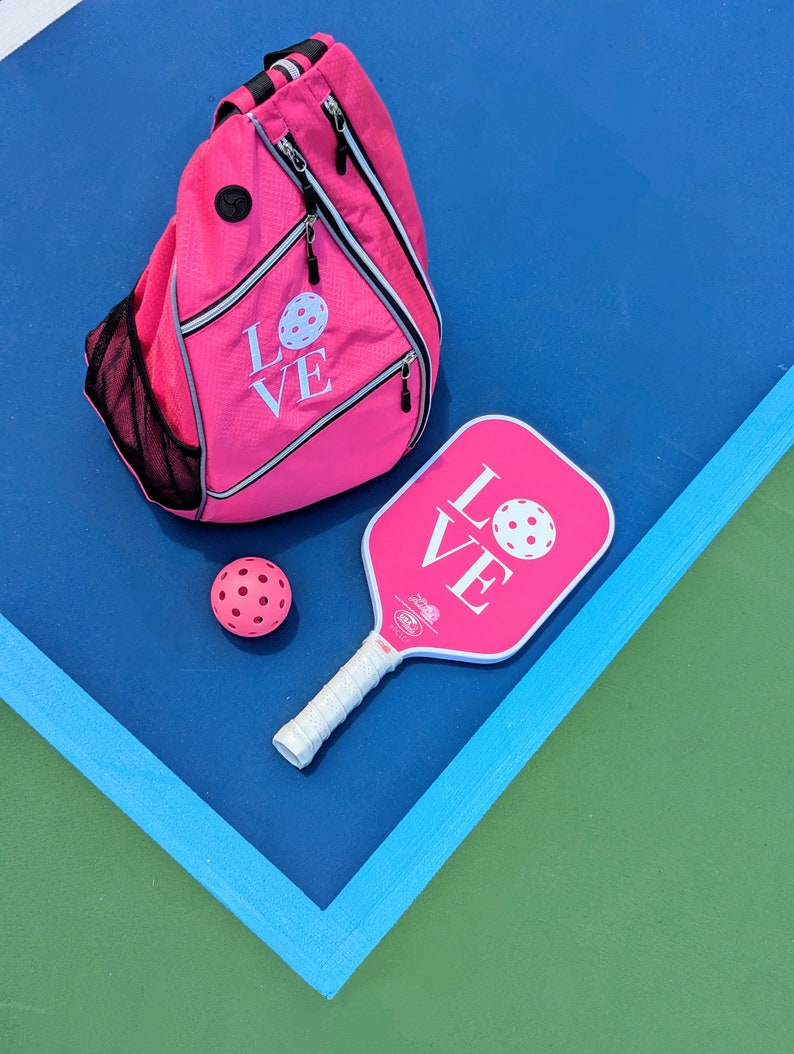 Puede incluir: Una pala de pickleball rosa con la palabra "LOVE" impresa en ella, una bolsa de pickleball rosa con la palabra "LOVE" impresa en ella y una pelota de pickleball rosa. La pala y la bolsa est&aacute;n sobre una superficie azul con una superficie verde en el fondo.