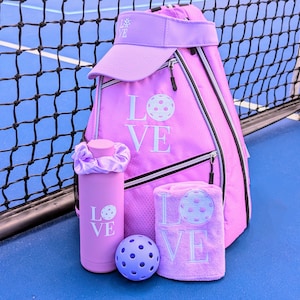 May include: A pink pickleball backpack with a matching pink visor, water bottle, towel, and ball. The backpack has the word "LOVE" embroidered on it with a pickleball graphic.