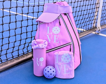 May include: A pink pickleball backpack with a matching pink visor, water bottle, towel, and ball. The backpack has the word "LOVE" embroidered on it with a pickleball graphic.
