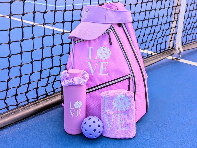 May include: A pink pickleball backpack with a matching pink visor, water bottle, towel, and ball. The backpack has the word "LOVE" embroidered on it with a pickleball graphic.