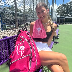 Puede incluir: Una mochila rosa con un gr&aacute;fico blanco de "LOVE" y un dise&ntilde;o de pickleball rosa. La mochila est&aacute; sentada en un banco morado.