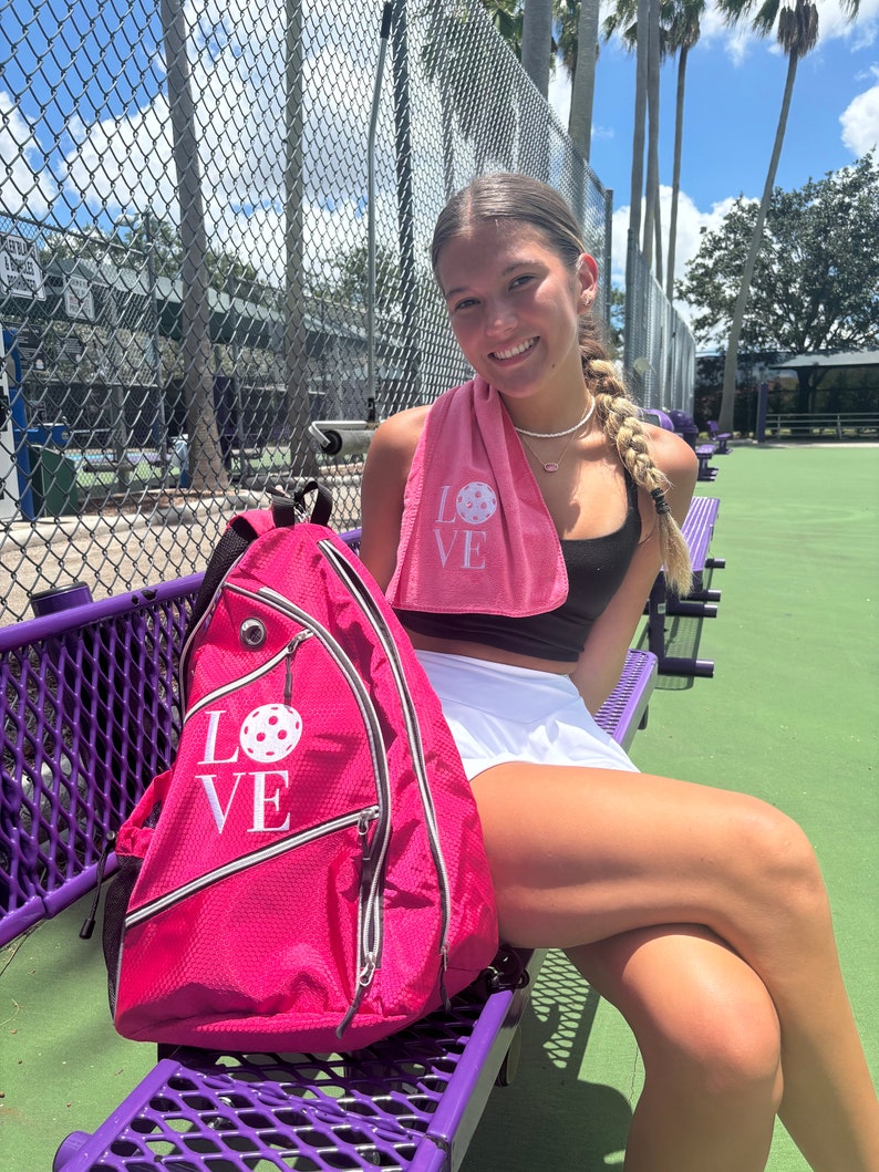 Puede incluir: Una mochila rosa con un gr&aacute;fico blanco de "LOVE" y un dise&ntilde;o de pickleball rosa. La mochila est&aacute; sentada en un banco morado.