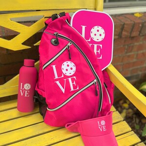 Puede incluir: Una pala de pickleball rosa, una mochila rosa con la palabra "LOVE" y un gr&aacute;fico de pickleball, una botella de agua rosa con la palabra "LOVE" y un gr&aacute;fico de pickleball, y una visera rosa con la palabra "LOVE" y un gr&aacute;fico de pickleball.