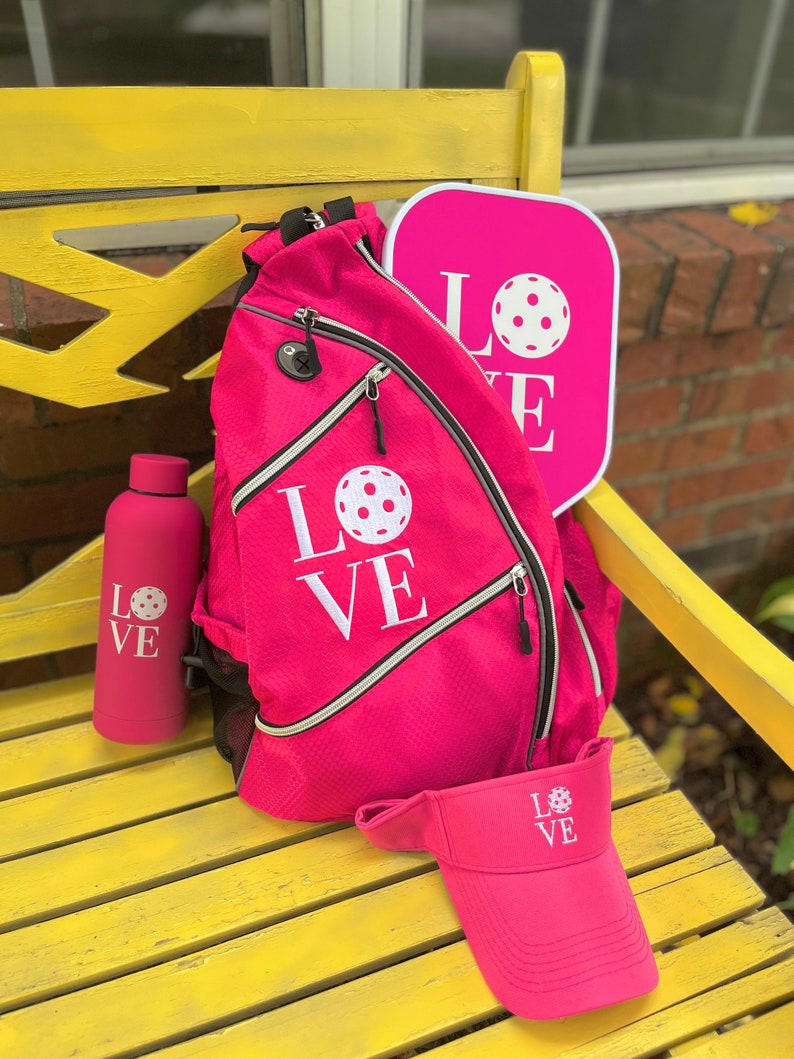 Puede incluir: Una pala de pickleball rosa, una mochila rosa con la palabra "LOVE" y un gr&aacute;fico de pickleball, una botella de agua rosa con la palabra "LOVE" y un gr&aacute;fico de pickleball, y una visera rosa con la palabra "LOVE" y un gr&aacute;fico de pickleball.