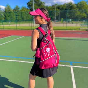 Puede incluir: Una mujer con una visera rosa y un vestido negro con una mochila rosa que dice "LOVE" con un gr&aacute;fico de pickleball blanco. Est&aacute; de pie en una cancha de pickleball verde.