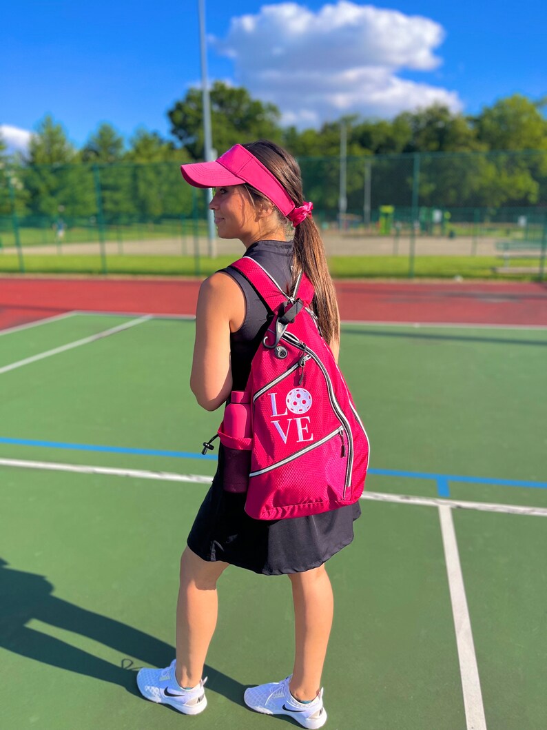 Puede incluir: Una mujer con una visera rosa y un vestido negro con una mochila rosa que dice "LOVE" con un gr&aacute;fico de pickleball blanco. Est&aacute; de pie en una cancha de pickleball verde.