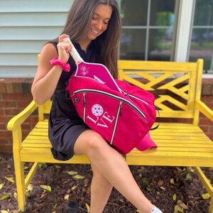 Puede incluir: Una mochila de pickleball rosa con la palabra "LOVE" bordada en la parte delantera. La mochila est&aacute; abierta y hay una pala de pickleball rosa dentro.