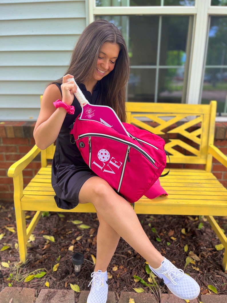 Puede incluir: Una mochila de pickleball rosa con la palabra "LOVE" bordada en la parte delantera. La mochila est&aacute; abierta y hay una pala de pickleball rosa dentro.
