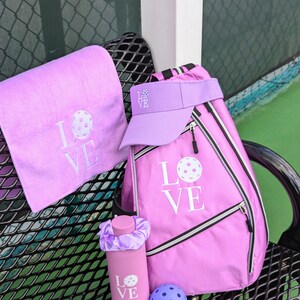 May include: A pink pickleball bag with a white "LOVE" design and a pickleball graphic. A pink water bottle with a white "LOVE" design and a pickleball graphic. A pink towel with a white "LOVE" design and a pickleball graphic. A pink visor with a white "LOVE" design and a pickleball graphic. A purple pickleball.