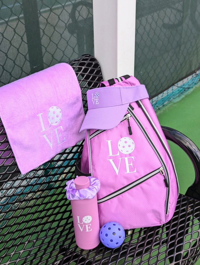May include: A pink pickleball bag with a white "LOVE" design and a pickleball graphic. A pink water bottle with a white "LOVE" design and a pickleball graphic. A pink towel with a white "LOVE" design and a pickleball graphic. A pink visor with a white "LOVE" design and a pickleball graphic. A purple pickleball.