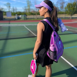 May include: A woman wearing a purple visor and a black sleeveless dress is holding a pink pickleball paddle. She is carrying a purple backpack with the word "LOVE" on it. The woman is standing on a pickleball court.