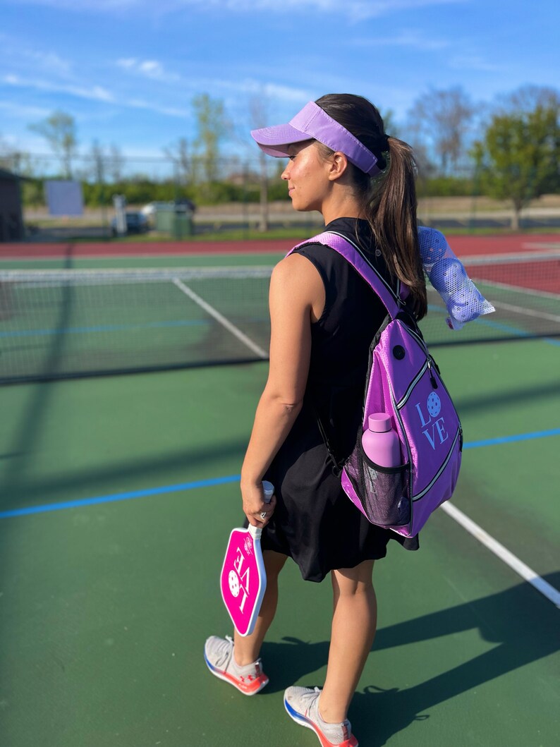 May include: A woman wearing a purple visor and a black sleeveless dress is holding a pink pickleball paddle. She is carrying a purple backpack with the word "LOVE" on it. The woman is standing on a pickleball court.