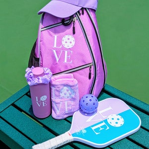 May include: A purple pickleball paddle with the word "LOVE" on it, a purple pickleball, a purple water bottle with the word "LOVE" on it, a purple towel with the word "LOVE" on it, and a purple pickleball backpack with the word "LOVE" on it.