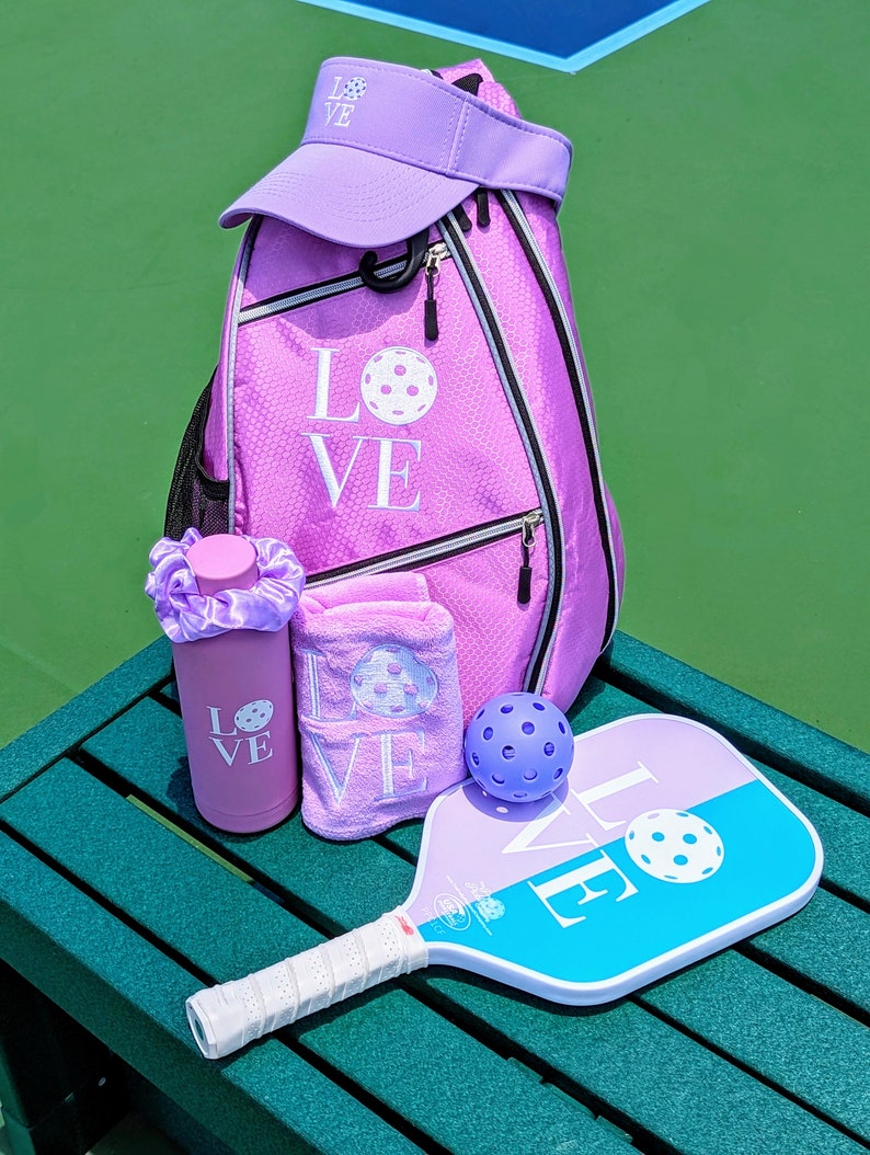 May include: A purple pickleball paddle with the word "LOVE" on it, a purple pickleball, a purple water bottle with the word "LOVE" on it, a purple towel with the word "LOVE" on it, and a purple pickleball backpack with the word "LOVE" on it.