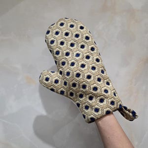 May include: An oven mitt with a beige and navy blue geometric pattern. The mitt is made of a textured fabric and has a loop for hanging. The design features a repeating pattern of navy blue circles within beige hexagons.