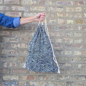 May include: A drawstring bag featuring a black and white chevron pattern. The bag is constructed from fabric and includes two white cord straps that cinch the top closed. The design consists of repeating zigzag lines in a high-contrast monochrome color scheme.