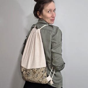 May include: A beige drawstring backpack with a patterned bottom section. The bag is made of a textured fabric and has a cream-colored drawstring closure. The bottom features a brown and gold ornate design.