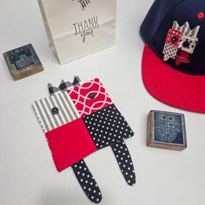 May include: A handmade fabric applique in red, black, and white patterns with button details. A navy blue and red baseball cap with a matching applique is also visible, along with two wooden stamps and a white gift bag with "Thank you" text.