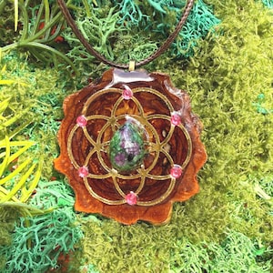 May include: A handcrafted pendant necklace featuring a brown, flower-shaped resin base with a gold-colored geometric design. The pendant has a central, oval-shaped stone with green and purple hues, and is accented with small pink gems. The necklace has a brown cord.
