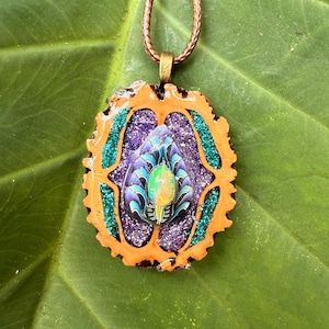 May include: A handcrafted pendant necklace featuring a unique design. The pendant has an orange border, teal accents, and a central design with purple glitter and a central iridescent stone. The necklace has a brown cord.