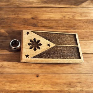 May include: A wooden plaque featuring the design of the Philippine flag, with a sun and three stars. The plaque is rectangular and has a natural wood finish. A silver ring sits next to the plaque.