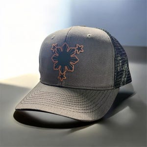 Op de afbeelding: Een grijze en zwarte truckercap met een koperkleurig geborduurd ontwerp van de Filippijnse zon en sterren.